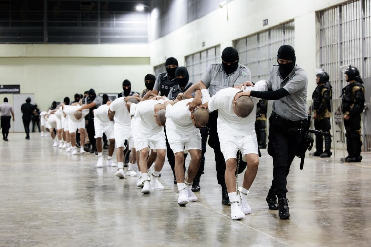 Men with shaved heads in white shorts and t-shirts being held down and marched along by prison guards wearing ski-mask type headgear.